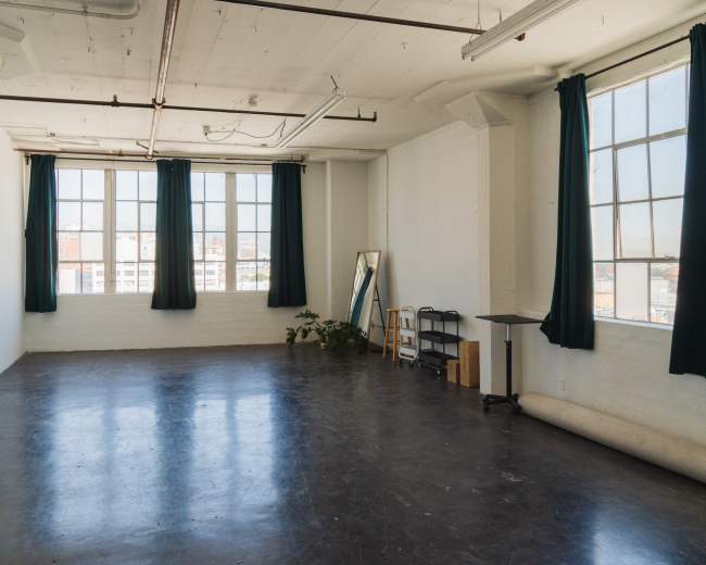 The image shows a spacious, sunlit room with large windows, green curtains, a mirror, a small table, and a roll of material on the floor.