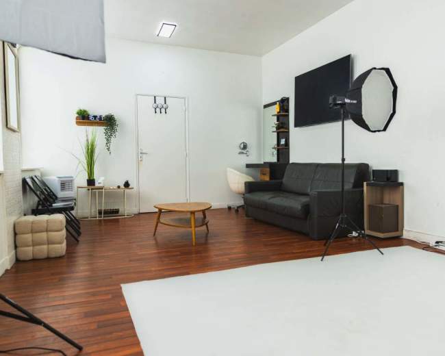 A spacious and well-lit photo studio features a black leather sofa, a wooden coffee table, and two softbox lights set up on tripods.