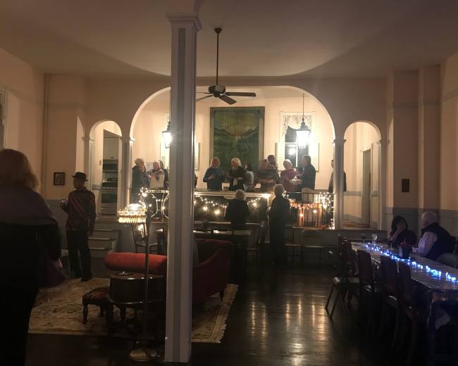 A group of people gathers on a balcony while others socialize in a warmly lit interior space with chairs and a rug.