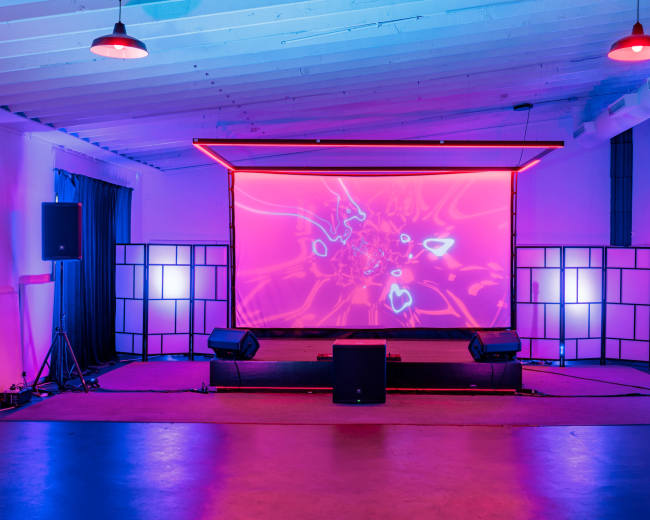 The image shows a stage setup with a large screen displaying colorful abstract visuals, flanked by speakers and illuminated by ambient lighting in a dimly lit room.