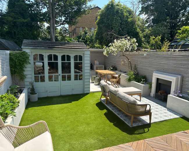 The image shows a landscaped garden featuring a lawn, outdoor seating area, a wooden dining table, and a small shed surrounded by greenery and a patio.