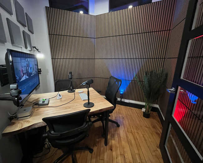 The image shows a small recording studio with wooden walls, a large monitor, two microphones on stands, and a minimalist wooden desk.