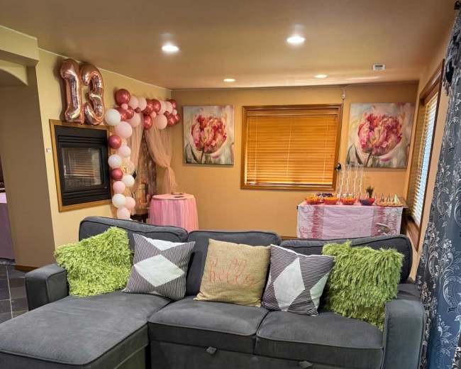 The image shows a living room with a gray sectional sofa adorned with decorative pillows, a large area rug, and a festive setup featuring a table decorated with balloons and flowers in the background.
