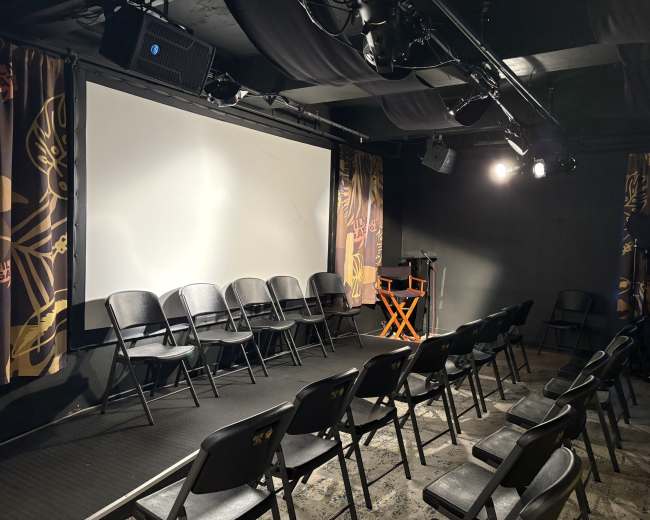 The image shows a small theater setup with rows of black chairs facing a large white screen.