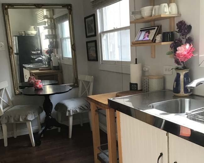 The image shows a small kitchen area featuring a table, two chairs, a mirrored wall, and a stainless steel sink with wooden cabinets.
