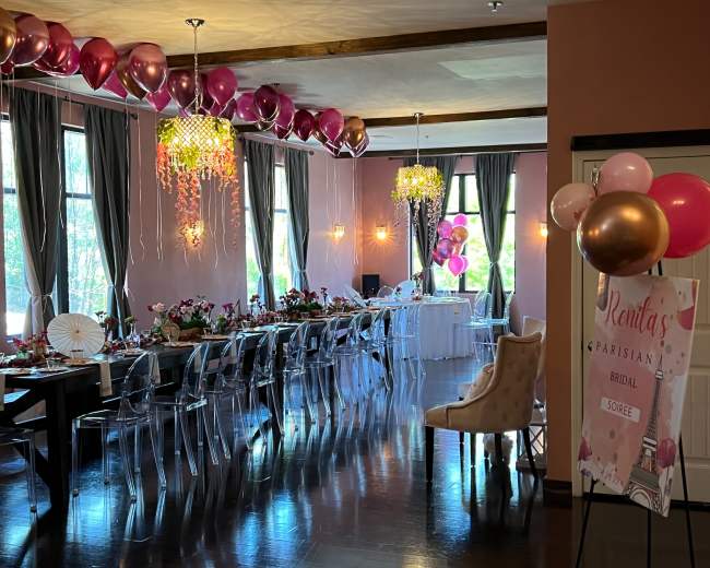 A decorated event space set up for a bridal shower, featuring a long table adorned with floral arrangements and pink balloons overhead.