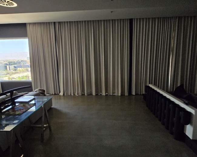 A spacious room features a large window covered by sheer curtains, a simple desk, and a sectional sofa against the wall.