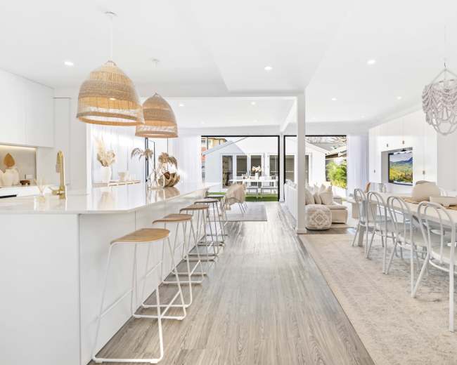 The image shows a bright, modern open-concept dining and living area with a white kitchen, wooden flooring, and ample natural light.