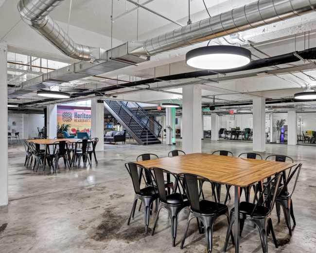 The image shows a spacious, modern office interior with exposed ceilings, multiple tables arranged for meetings, and glass-walled conference rooms in the background.