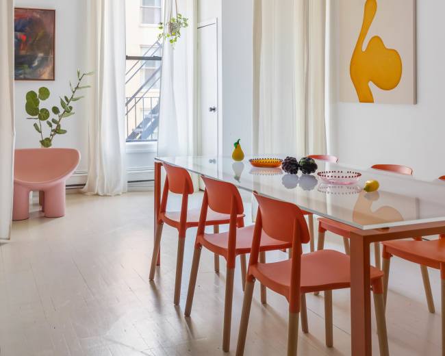 A bright dining area features a white table surrounded by orange chairs, with curtains, artwork, and decorative objects enhancing the modern aesthetic.