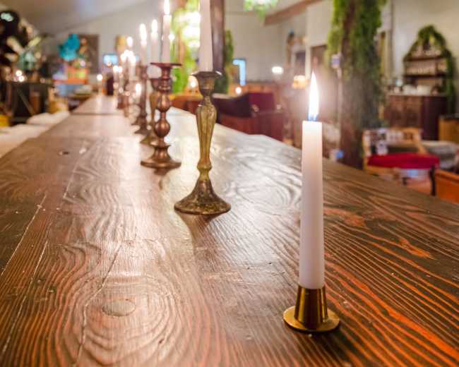 The image shows a wooden table with several candlesticks and flickering candles, set in a warmly decorated indoor space adorned with greenery.