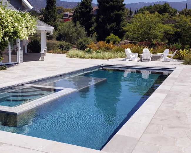 The image shows a modern stone patio with a rectangular swimming pool and a seating area featuring white chairs and a table amidst a landscaped yard.