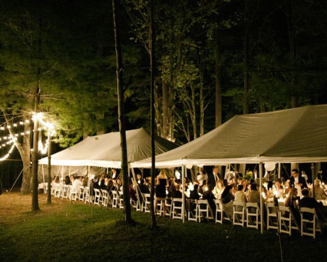 A group of people gather under string lights at tables arranged beneath large tents in a forested setting at night.