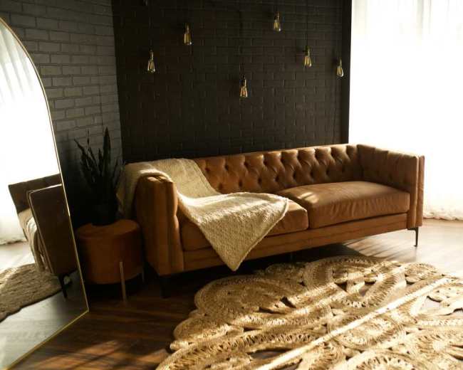 The image shows a cozy living room featuring a tan leather sofa, a large round woven rug, and natural light streaming through sheer curtains.