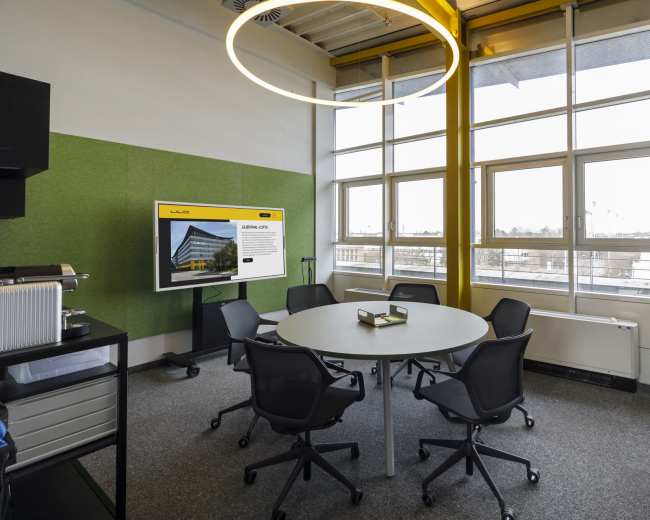 The image shows a modern conference room with a round table surrounded by six chairs, a large screen displaying information, and large windows letting in natural light.