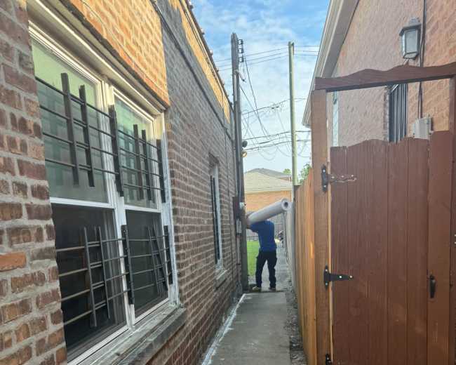 A person is standing in a narrow alleyway between two buildings, holding a rolled-up object near a wooden fence.