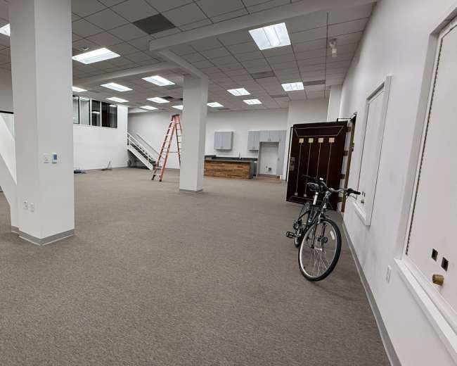 The image shows a spacious, empty office interior with beige carpeting, large windows, a kitchen area, a ladder, and a bicycle parked against the wall.