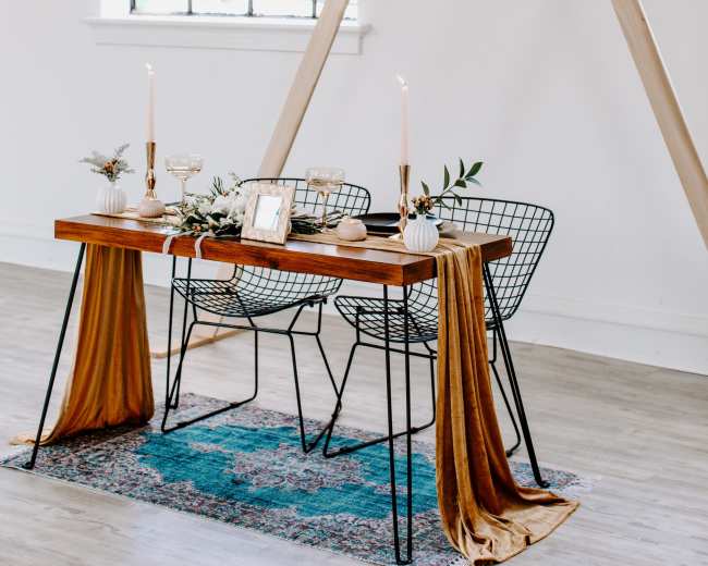 A wooden table with two wire chairs is set up in front of a large window, adorned with candles, decorative items, and a triangular wooden structure overhead.