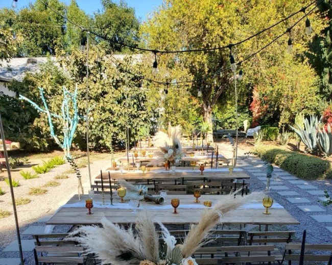 The image shows a beautifully arranged outdoor dining setup with long wooden tables, adorned with floral centerpieces and decorative candles, in a garden setting.