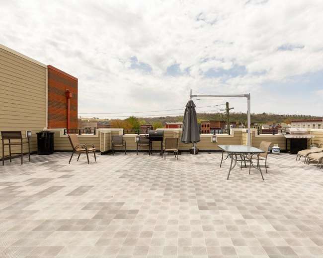 The image shows a spacious rooftop terrace with chairs, tables, and a large umbrella against a backdrop of buildings and a cloudy sky.
