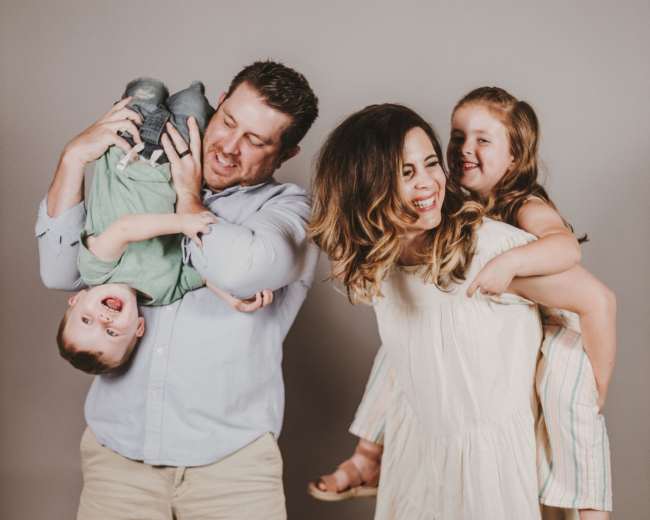 A family poses for a photo, with the father holding a boy upside down and the mother carrying a girl on her back.