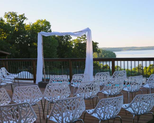 The image shows a wedding ceremony setup with white chairs arranged in rows beneath a white draped arch overlooking a lake.