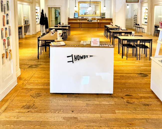 The image shows a spacious retail store with wooden flooring, featuring a central table surrounded by black chairs and shelves displaying various products along the walls.