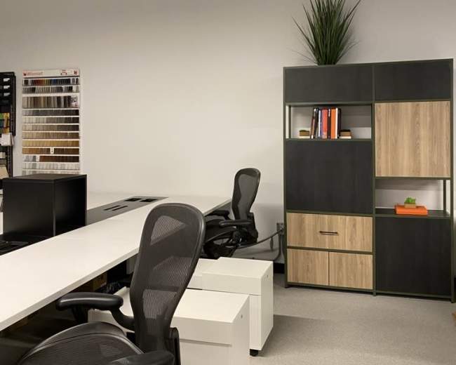 The image shows a minimalistic office space with two black ergonomic chairs and a long white desk, accompanied by a storage unit featuring shelves and drawers.