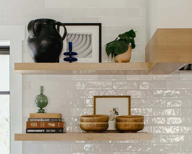 The image features a kitchen with two floating shelves displaying decorative items, including books, bowls, and a potted plant, above a countertop with a cutting board and lemons.