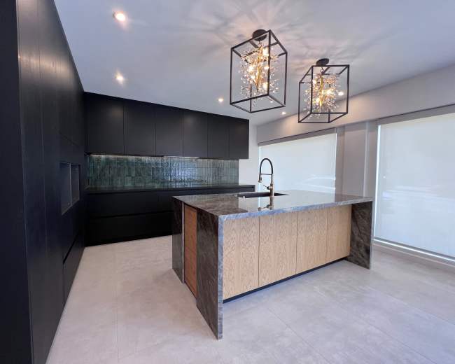 The image shows a modern kitchen featuring a large island with a mix of dark stone and light wood, black cabinetry, and unique pendant lighting.