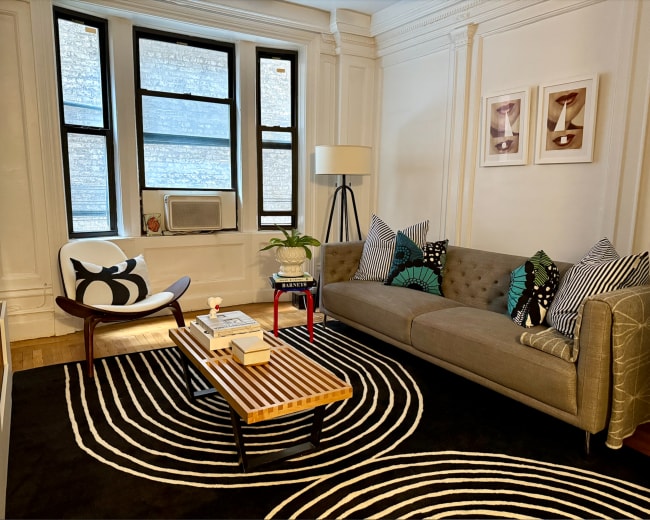 The living room features a gray tufted sofa, a striped area rug, a round coffee table, and large windows that provide natural light.