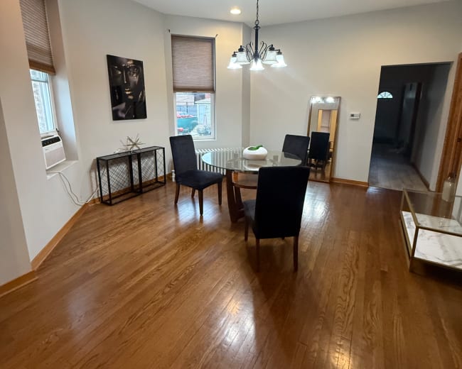 A dining area features a round table with four chairs, a chandelier, and hardwood flooring, alongside a wall with window shades and a mirror.