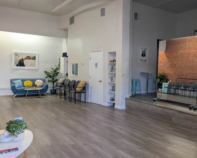 The image shows a spacious interior with a seating area featuring blue and yellow decor on one side, and a small stage with chairs arranged in rows on the other side, set against a brick wall.
