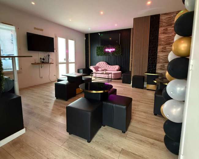 The image shows a modern lounge area featuring black seating, a pink sofa, a television on the wall, and decorative balloons along one side.