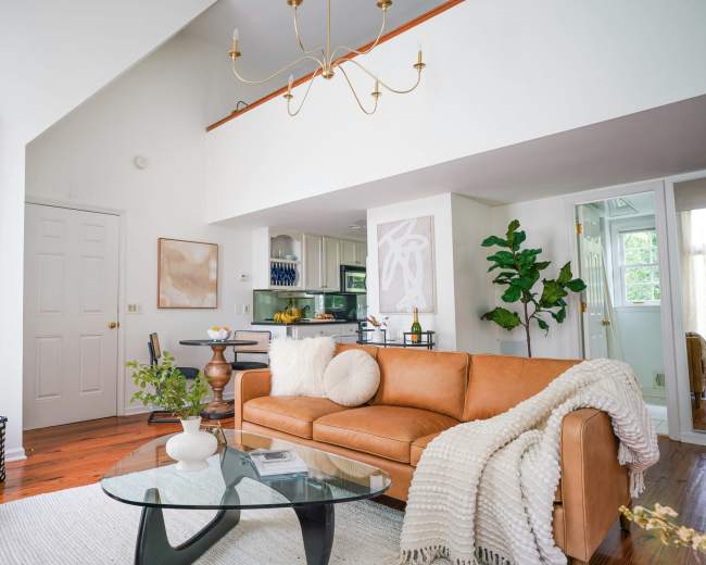 A bright living room features a tan leather sofa, a glass coffee table, and a kitchen area visible in the background.
