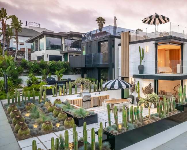 A modern two-story house with large windows is surrounded by a landscaped area featuring various cacti and a barbecue setup, topped with a black and white striped umbrella.