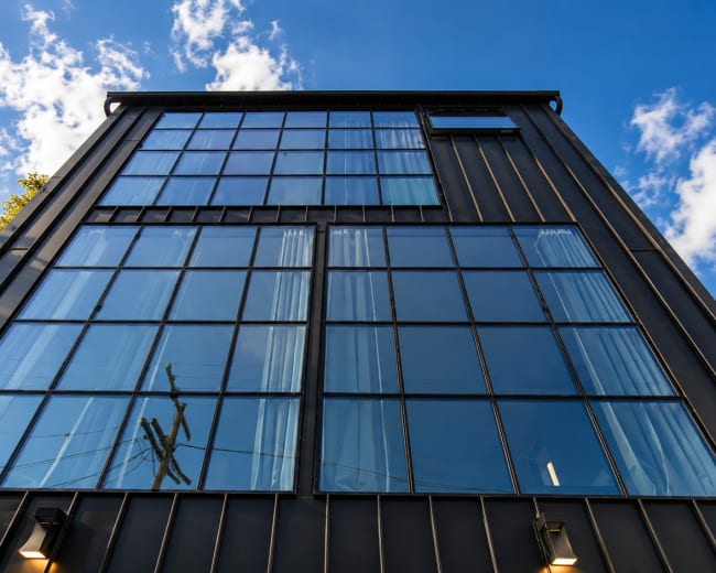 The image shows a modern building with a black metal facade and large glass windows reflecting the sky and surrounding trees.