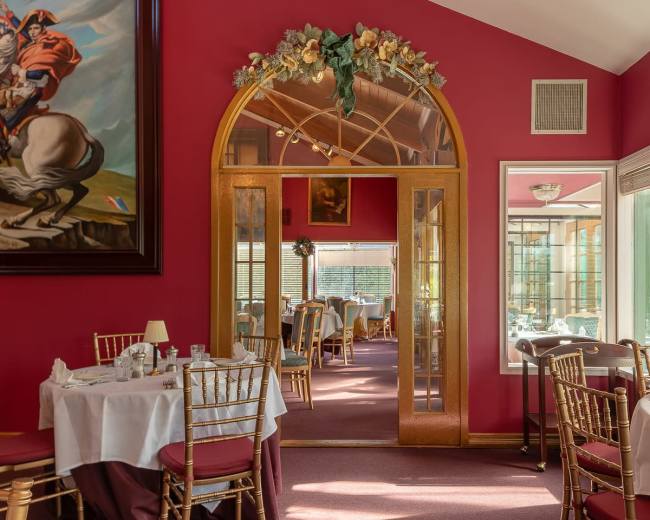 The image shows a dining area with red walls, elegant round tables, and a large mirror reflecting a decorative archway.