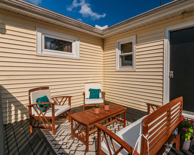 A small outdoor patio features two wooden chairs and a table, surrounded by yellow siding and a single door.