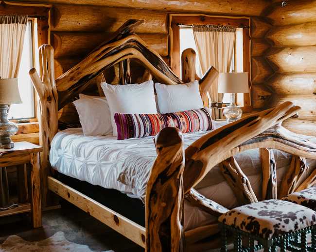 A rustic log cabin bedroom featuring a wooden bed frame with intricate branch-like details, a white bedspread, and two bedside lamps.