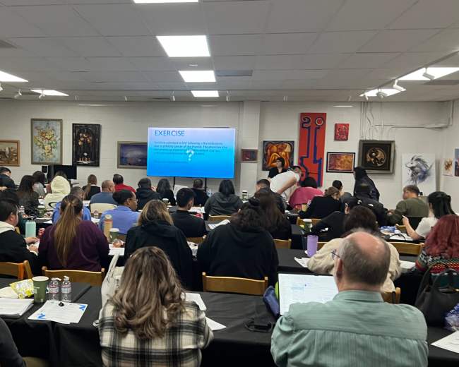 A classroom filled with students seated at tables, attentively working on exercises while facing a presentation screen displaying instructions.