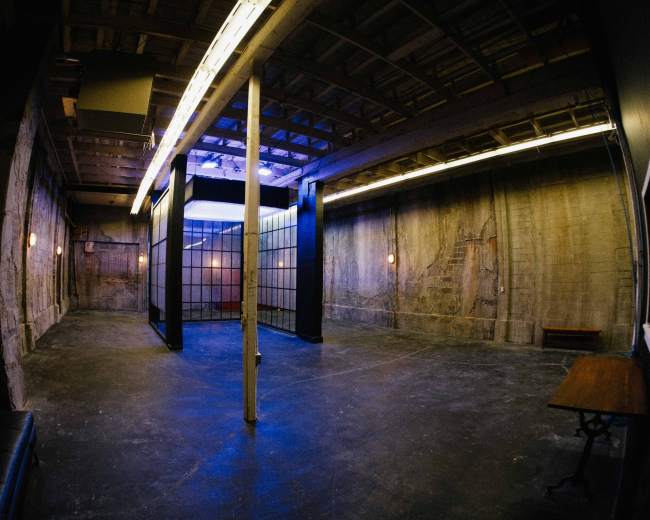 The image depicts an empty, industrial-style room with concrete walls, a grid partition, and sparse lighting.