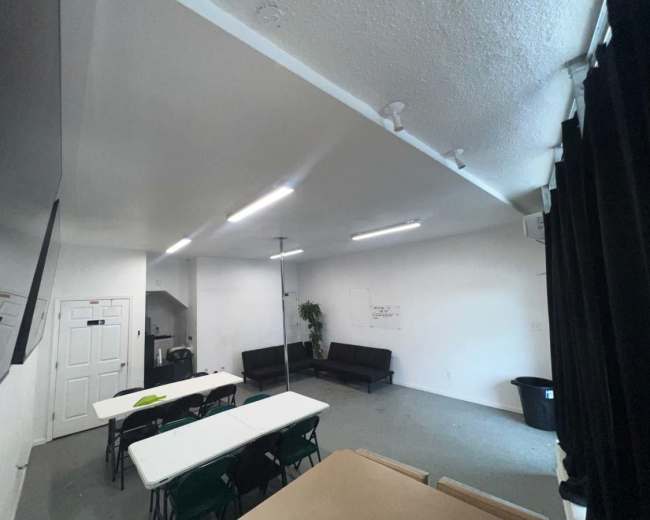 The image shows a spacious room with a few tables, chairs, a black couch, and a small kitchenette area.