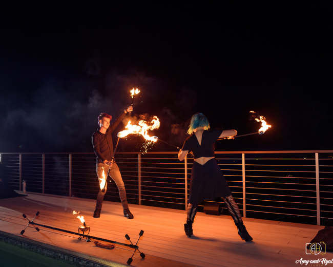 Two performers with flaming staffs dance on a wooden deck illuminated by lights at night, while a seated person watches in the background.