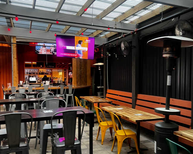 The image shows a partially covered outdoor seating area with metal and wooden tables, colorful chairs, and a bar in the background, illuminated by overhead lights and a screen.