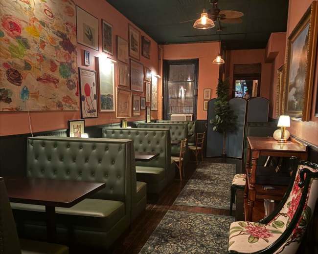The image depicts a narrow restaurant interior with green upholstered booths along the walls, framed artwork, and warm lighting.