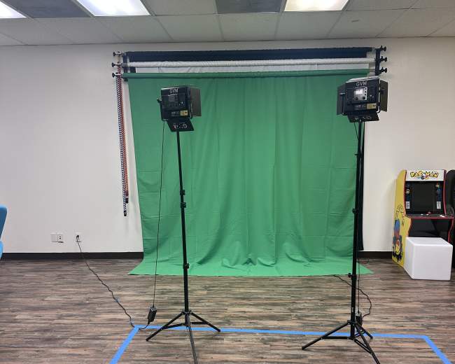 A room with a green screen setup flanked by two lighting stands, and an arcade machine in the background.