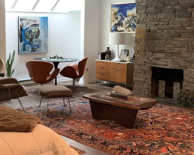 A modern living space with a bed in the foreground, a stone fireplace, a wooden coffee table, and a dining area featuring a round table and chairs.