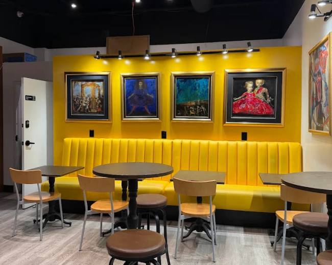 The image shows a cafe interior with yellow upholstered seating, round tables, and framed paintings on the wall.