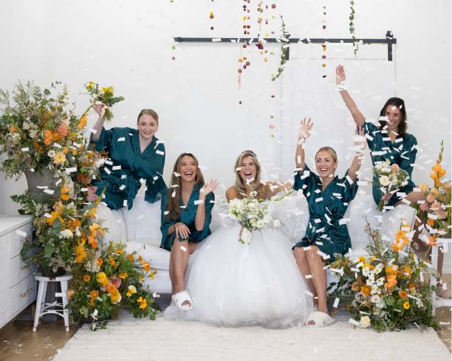 A group of five women in green outfits celebrate around a bride in a white dress, with flowers and petals falling in a bright, decorated space.